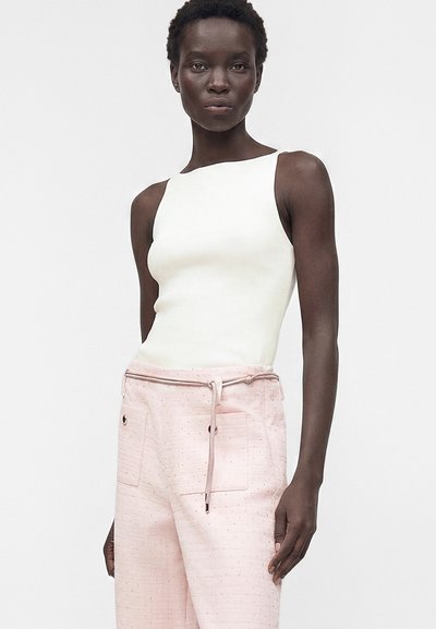 Femme aux cheveux courts porte un haut blanc sans manches et un pantalon rose pâle taille haute avec poches et ceinture à cordon.