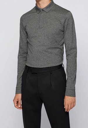 Mand iført en tætsiddende grå langærmet polo t-shirt stoppet ned i sorte skræddersyede bukser foran en ensfarvet baggrund.