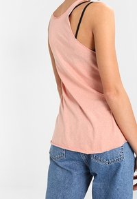 Ljusrosa linne med avslappnad passform, med en rundad halsringning och en böjd nederkant. Svarta band är synliga under linnet.