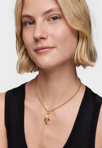 Collier en perles dorées avec un pendentif en forme d'ours en peluche, présentant une texture lisse et une finition polie, porté sur un haut noir.