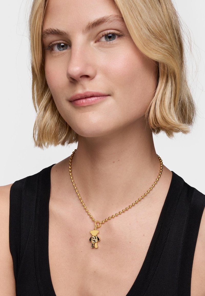 Collier en perles dorées avec un pendentif en forme d'ours en peluche, présentant une texture lisse et une finition polie, porté sur un haut noir.