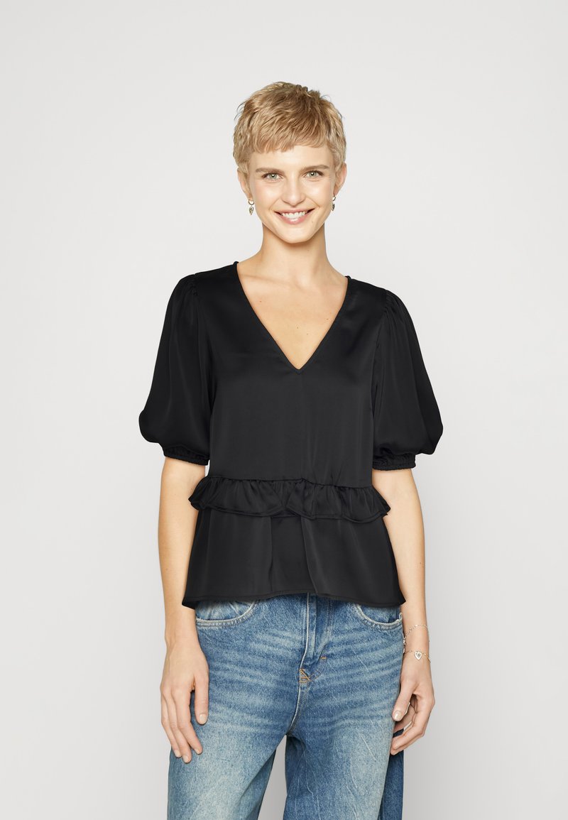 Blusa nera a maniche corte con scollatura a V e orlo peplum arricciato, realizzata in tessuto morbido. Indossata con jeans in denim azzurro chiaro.