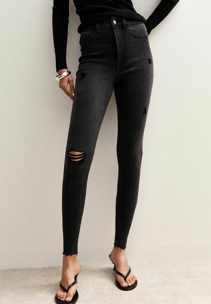 Jeans skinny de talle alto en negro con detalles desgastados, que presentan dos desgastes en las rodillas y dobladillo sin rematar, acompañados de chanclas negras.