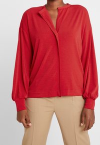 Blusa rossa a maniche lunghe con scollatura a V, vestibilità comoda e polsini arricciati. Tessuto morbido con una texture liscia; nessun motivo o accessorio visibile.