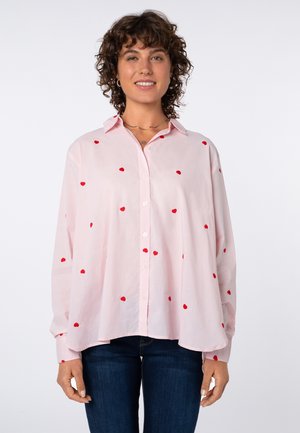 Frau mit lockigem Haar, die eine hellrosa Bluse mit roten Polka-Dots trägt, kombiniert mit dunkelblauen Jeans, steht vor einem einfarbigen Hintergrund.
