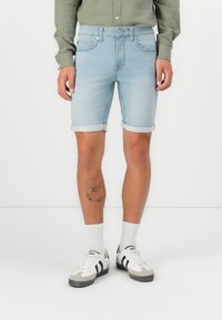 Pantalones cortos de denim azul claro con dobladillo vuelto, cinco bolsillos y un diseño ajustado. Llevados con calcetines blancos y zapatillas con acentos negros.