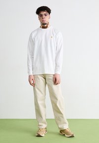 Chemise blanche à manches longues avec un petit logo, pantalon beige, baskets beige et marron, et lunettes de soleil rondes lilas claires, sur un sol vert.