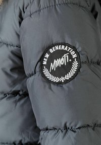 Grå puffjacka med en strukturerad yta och en rund svart lappar. Lappen har vit text och en lagerkransdesign, märkt "NYA GENERATIONEN."