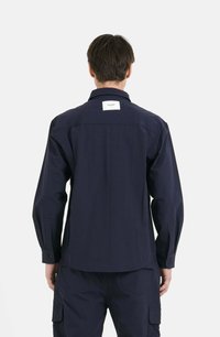 Camicia a maniche lunghe blu navy con colletto a bottoni, yoke posteriore e etichetta del marchio ben visibile. Realizzata in un tessuto liscio e resistente, con una vestibilità comoda.