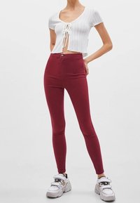 Mujer vestida con pantalones skinny de cintura alta burdeos, blusa blanca corta de manga con nudos en el frente y zapatillas blancas gruesas, de pie frente a un fondo simple.