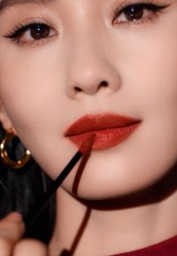 Flüssiger Lippenstift wird auf volle Lippen mit einem matten, tiefroten Finish aufgetragen. Das Modell trägt Kreolen und hat eine glatte, helle Haut.