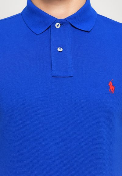 Zila polo krekls ar divām baltām pogām un mazu sarkanu izšūtu polo spēlētāja logo uz krūšu, ko valkā cilvēks.
