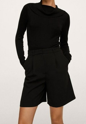 Personne portant un haut noir à manches longues et un short noir taille haute, les mains dans les poches, sur un fond uni.