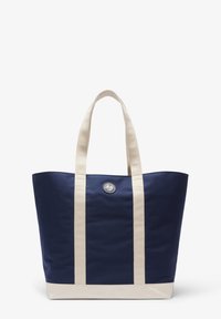 Sac fourre-tout bleu marine avec poignées et base beige. Présente un écusson logo et un design structuré. Fabriqué en tissu durable à la texture lisse.