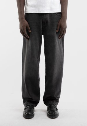 Osoba stojąca, ubrana w czarne dżinsy o szerokich nogawkach, czarne skórzane buty i białą koszulę, widziana od połowy tułowia do stóp.