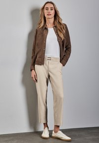 Brun mockaskinnjacka över en vit t-shirt, beige skräddarsydda byxor och vita sneakers med en tan-färgad sula, mot en neutral bakgrund.