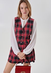 Abito a vestito a quadri rossi e neri con una camicia bianca a maniche lunghe. Gli accessori includono una borsa a tracolla in pelle di coccodrillo rossa con hardware dorato.