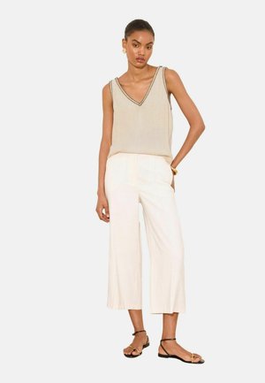 Vrouw staat en draagt een beige mouwloos V-hals topje, witte wijde cropped broek, zwarte sandalen met enkelbandje en gouden sieraden.