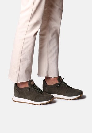 Sneakers scuri in suede verde con superficie texturizzata, suole bianche e lacci verdi, abbinati a pantaloni cropped beige chiaro con orlo sfrangiato.