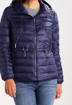 Frau trägt eine marineblaue gesteppte Jacke mit Reißverschluss vorne und Reißverschlusstaschen an den Seiten, kombiniert mit blauen Jeans und einem grauen Oberteil darunter.