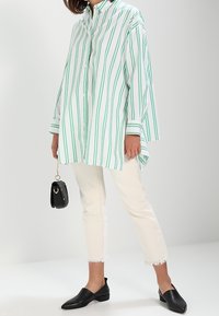 Žena v volné bílé košili se zelenými vertikálními pruhy, bílých zkrácených kalhotách, černých nazouvacích botách, držící malou černou kabelku.