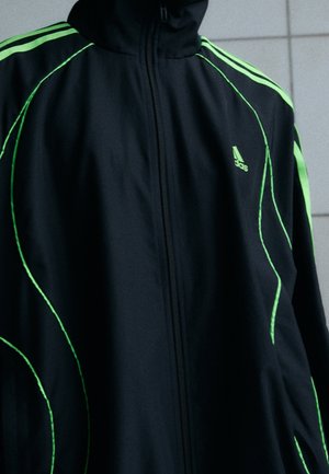 Zwarte Adidas-jas met neon groene strepen en gebogen lijnen, voorste rits gesloten, gedragen door een persoon tegen een betegelde muur.
