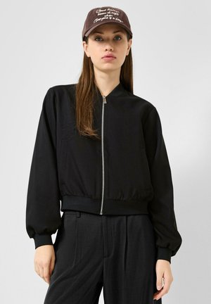 Frau trägt eine schwarze Zip-Jacke, dunkle Hose und eine braune Kappe mit weißem Schriftzug, steht vor einem schlichten hellen Hintergrund.