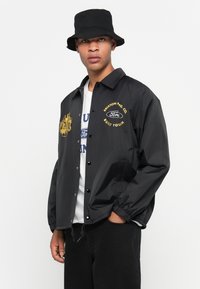 Black lightweight jacket with yellow embroidery, button closure, and elastic cuffs. Paired with a white t-shirt and black bucket hat.