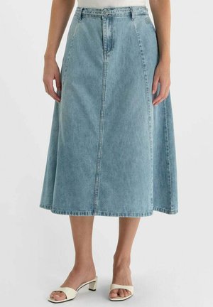 Vrouw met een lichtblauwe denim midirok met hoge taille en witte open-toe sandaaltjes met hak, staand tegen een effen achtergrond.