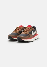 Brown and black athletic shoes with a white midsole, red accents, grey detailing, and a textured rubber outsole. Features laces and a swoosh logo.