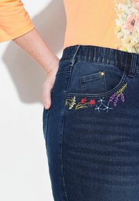 Czarna dżinsowa spódnica z elastycznym paskiem, z wielokolorowym haftem kwiatowym na prawej kieszeni. Gładka tekstura, dopasowany krój.