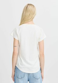 T-shirt bianca in cotone con maniche corte e scollo rotondo, caratterizzata da un orlo curvo e una texture liscia, abbinata a jeans azzurri chiari.
