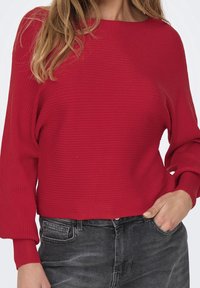 Pull rouge côtelé à large encolure et manches longues, associé à un jean gris foncé. Le tissu a une surface texturée.