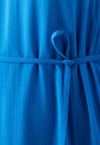 Blå kjole med strukturert stoff og prikkete mønster, knyttet i midjen med et matchende belte. Lett drapert design.