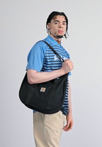 Sac en toile noir avec un patch logo Carhartt, de forme arrondie et avec des bandoulières robustes, porté de manière décontractée sur l'épaule.