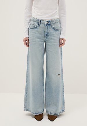 Persoon draagt lichtblauwe jeans met wijde pijpen en kleine scheurtjes op de dij en knie, gecombineerd met een wit shirt met lange mouwen en bruine puntige schoenen.