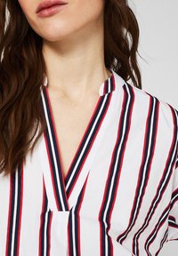 Blusa bianca a righe con strisce verticali rosse e blu navy, con scollatura a V e una vestibilità comoda. Tessuto leggero e morbido con una leggera lucentezza.