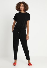 Zwart jumpsuit met korte mouwen en een elastische trekkoord taille. Zachte stof, taps toelopende broekspijpen, en geaccentueerd met rode sandalen.