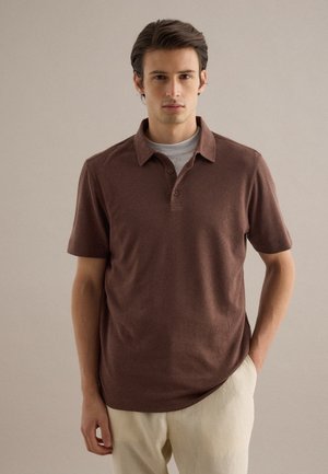 Uomo che indossa una polo marrone a maniche corte sopra una maglietta grigia e pantaloni beige, in piedi davanti a uno sfondo semplice di colore grigio chiaro.