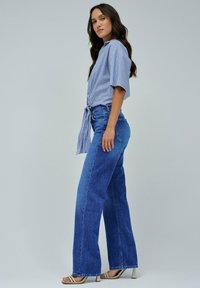 Camicetta a righe blu a maniche corte annodata in vita, abbinata a jeans a gamba larga blu e sandali con tacco di colore chiaro. Design semplice.