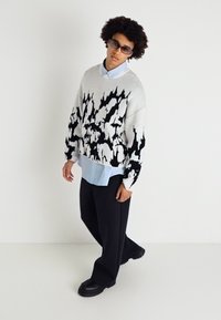 Pull surdimensionné gris avec des motifs abstraits noirs, superposé sur une chemise col bleu clair, associé à un pantalon large noir et des chaussures épaisses.