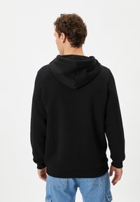 Zwarte hoodie met een grote capuchon, geribbelde manchetten en zoom, gemaakt van zachte stof. Draagt een lichte blauwe cargobroek. Geen zichtbare logo's.