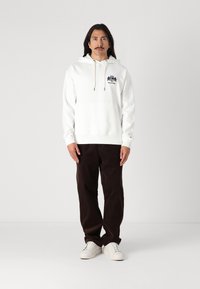 Weißer Kapuzenpullover mit vorderer Kängurutasche, Zugbandverschluss und gesticktem Logo. Kombiniert mit dunkelbraunen Cordhosen und weißen Sneakers.