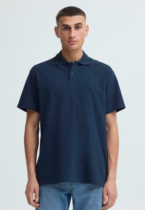 Muž v tmavě modrém krátkém polokošili a světle modrých džínách stojí před jednobarevným světlým pozadím.