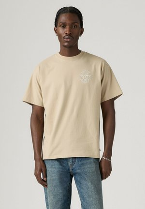 Młody mężczyzna z warkoczami, ubrany w beżowy t-shirt "Sun Seeker" i niebieskie dżinsy, stojący na jednolitym jasnym tle.