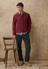 Burgunderfarbener Fleece-Cardigan mit Knopfverschluss und zwei vorderen Taschen, getragen über einem blauen Jeanshemd, kombiniert mit dunklen Hosen und hellbraunen Schuhen.