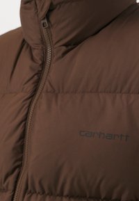 Braune Pufferjacke mit Reißverschluss, ausgestattet mit einem gesteppten Design und dem "Carhartt"-Logo in Dunkelblau auf dem Stoff.