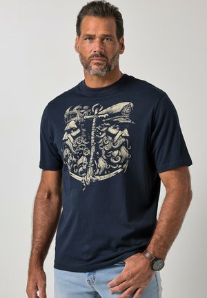 Homme barbu portant un t-shirt bleu marine affichant un graphisme crème détaillé de deux visages barbus et une ancre, associé à un jean bleu clair et une montre.