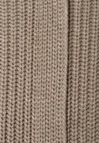 Beige stickad tyg med ribbad textur, med rader av jämnt fördelade stygn som skapar ett konsekvent mönster.