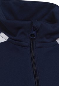 Veste zippée bleu marine avec un col haut, présentant une texture de tissu lisse et une bande blanche contrastante sur l'épaule.